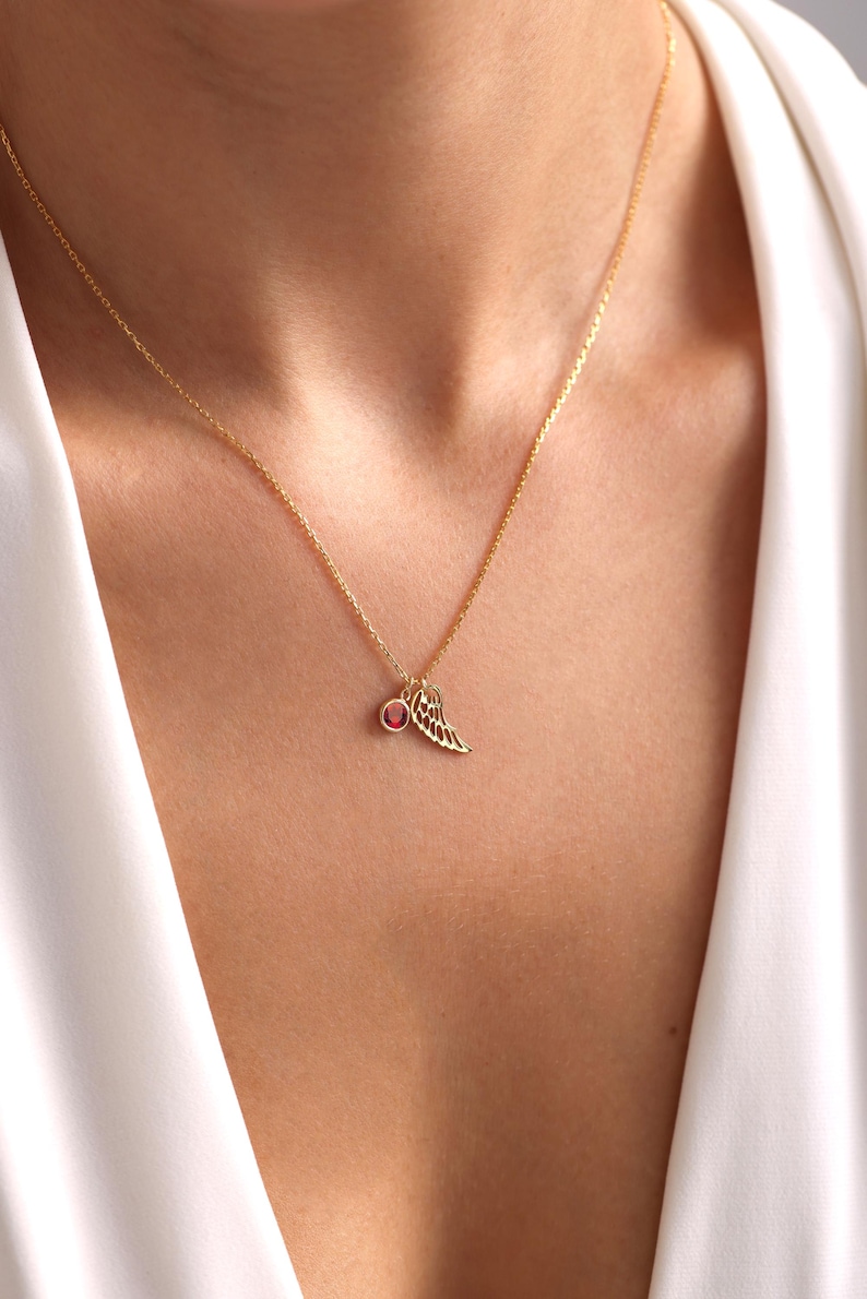 Op de afbeelding: Een gouden ketting met een kleine rode edelsteen en een delicate gouden vleugelbedel.