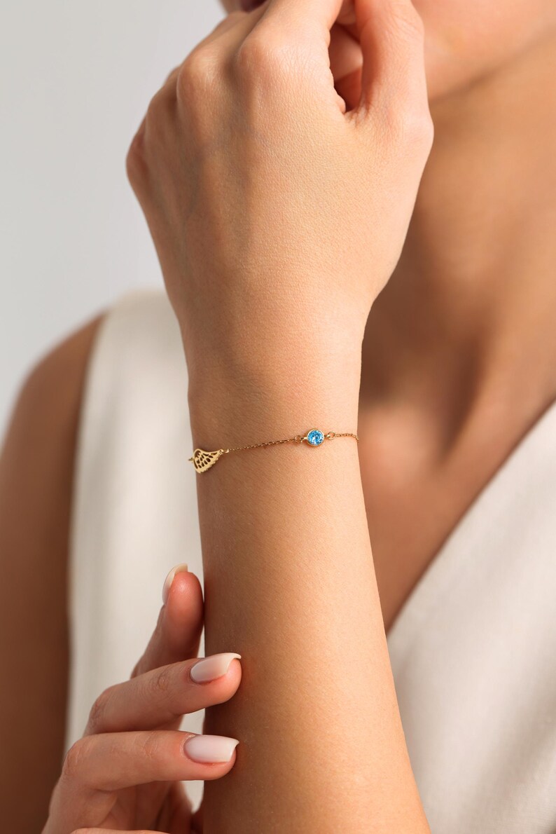 May include: A delicate gold bracelet with a small, openwork wing charm and a round, light blue gemstone. The bracelet is worn on a wrist, highlighting its simple yet elegant design. The jewellery is a subtle accessory.