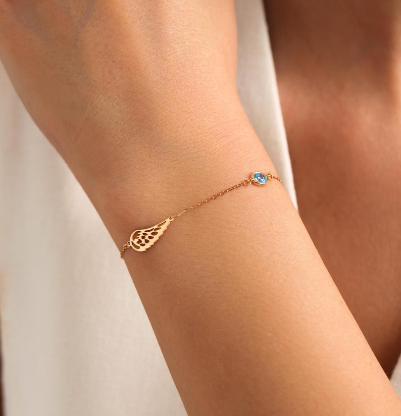 May include: A gold bracelet with a wing-shaped charm and a small, round, light blue gemstone. The bracelet is attached to a fine chain and is worn on a wrist.