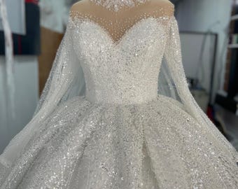 Puede incluir: Un vestido de novia blanco con mangas largas y cuello alto. El vestido está cubierto de lentejuelas y tiene una falda completa.