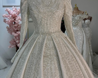 Puede incluir: Un vestido de novia blanco con mangas largas y cuello alto. El vestido está cubierto de lentejuelas y tiene un corpiño ajustado y una falda completa.