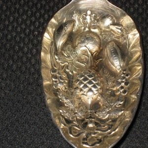 Puede incluir: Una cuchara de plata con un diseño ornamentado que presenta una piña, uvas y hojas. La cuchara es muy detallada y tiene una estética vintage.
