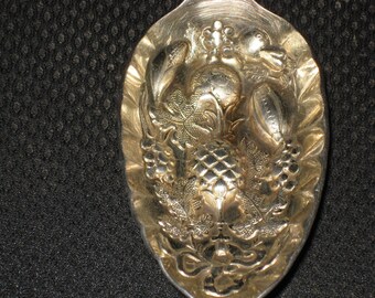May include: A silver spoon with an ornate design featuring a pineapple, grapes, and leaves. The spoon is highly detailed and has a vintage aesthetic.