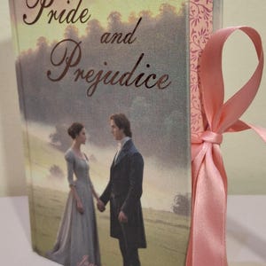 May include: A hardback copy of "Pride and Prejudice" featuring a romantic scene on the cover. The book is tied with a pink ribbon. The author's name, Jane Austen, is printed on the cover. The design has a vintage, soft colour palette.