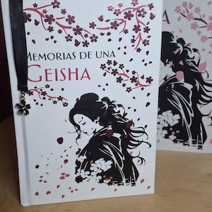 May include: Two white hardcover books with pink and white cherry blossom designs. The book on the left has the title "Memorias de una Geisha" and a black ribbon bookmark. The book on the right has the text "De una" visible.