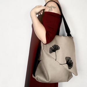 Può includere: Borsa a tracolla beige con cinturino nero e chiusura con patta. La borsa presenta due disegni di foglie di ginkgo nere. La persona nell'immagine indossa un vestito bordeaux.