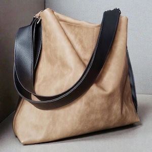 May include: A tan-colored tote bag with a wide black shoulder strap. The bag has a soft, slouchy design and a contrasting black strap. The bag appears to be made of leather or a leather-like material.