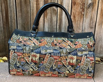 May include: A black leather-trimmed travel bag with a tapestry-like design featuring a colorful pattern of travel-themed words and logos, including "PAN AM", "DELTA", and "SWISS".