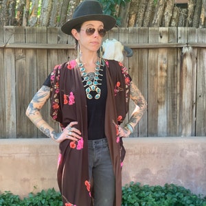 May include: A woman wearing a brown floral print kimono with a black top and dark wash jeans. She is wearing a large turquoise and silver statement necklace and a black fedora hat.