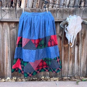 May include: A blue denim skirt with a patchwork design. The skirt is made of different fabrics, including plaid and solid colors. The skirt is hanging on a wooden fence.