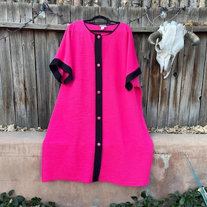May include: A bright pink dress with black trim and buttons. The dress has short sleeves and a textured fabric. It is hanging on a black hanger against a wooden fence background.