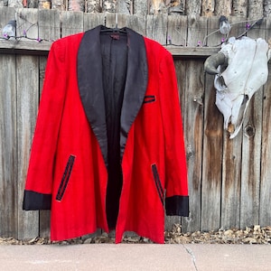 May include: A vintage red blazer with a black satin shawl collar and black accents on the pockets and cuffs. The jacket is hanging on a wooden fence. The blazer is a classic piece of outerwear.