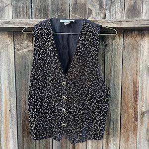 May include: A black velvet vest with a speckled pattern and gold-colored buttons. The vest has a V-neck and is hanging on a gold-colored hanger against a wooden background. The vest is buttoned up.