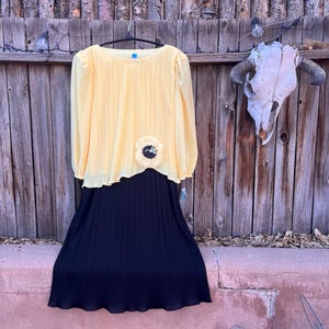 May include: A two-piece dress with a yellow pleated top and a navy blue pleated skirt. The top has long sleeves and a decorative flower. The dress is hanging on a black hanger against a wooden fence.