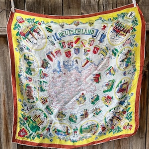 May include: A square scarf featuring a map of Germany with the word "Deutschland" at the top. The scarf has a white background with a yellow border and a brown outer edge. It includes illustrations of landmarks and coats of arms.