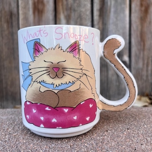 May include: White ceramic mug with a cat illustration. The cat is light brown with a pink nose and is resting on a pink pillow with white hearts. The mug has a tail-shaped handle and the text "What's Snooze?" in pink.