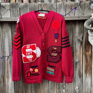 May include: A red vintage cardigan sweater with various patches and appliqués. Features include a smiley face, the letter "S", the word "Donna", and the text "CAPT 72". The sweater has black stripes and a purple ribbon.