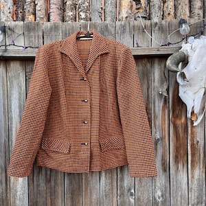 May include: A vintage houndstooth blazer in shades of orange and black hangs on a wooden fence. The blazer features a classic collar, button closure, and two front pockets. The background includes a rustic wooden fence and a cow skull.