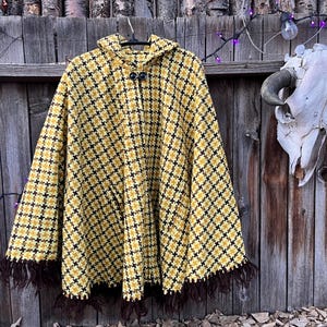 May include: A hooded poncho in a yellow and black houndstooth pattern, with brown fringe along the bottom. The poncho has two black buttons and is hanging on a wooden fence. A skull with horns is mounted on the fence.