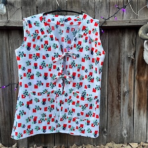 May include: A sleeveless white vest with a red and blue floral pattern. The vest has two pockets and ties in the front. The vest is hanging on a black hanger against a wooden background.