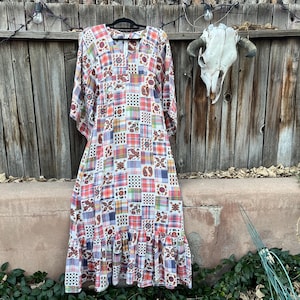 May include: A long, patchwork-style dress with a ruffled hem, hanging on a black hanger. The dress features a mix of plaid and paisley patterns in red, orange, blue, and white. The background includes a wooden fence and a cow skull.