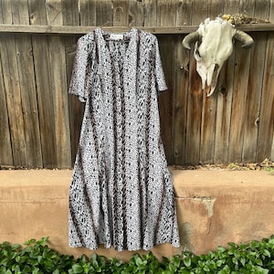 May include: A long, flowing dress with a black and white pattern hangs on a wooden fence. The dress has short sleeves and a V-neck. The pattern is a vertical design with brown accents. A cow skull decorates the fence.
