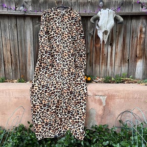 May include: A long-sleeved, floor-length robe featuring a bold leopard print pattern in shades of black, brown, and tan. The garment has a full-length front button closure, a rounded neckline, and a loose, straight-cut silhouette throughout.