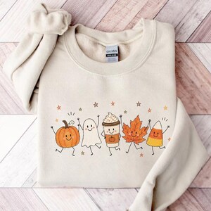 May include: A sand-colored crewneck sweatshirt featuring a graphic print of five smiling autumn-themed characters: a pumpkin, a ghost, a coffee cup, a maple leaf, and a piece of candy corn, all surrounded by small star shapes.