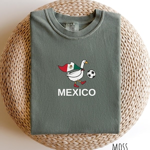 May include: A sage green crewneck t-shirt featuring a graphic of a white goose wearing red shoes and carrying a Mexican flag. The goose is positioned next to a black and white soccer ball. The word MEXICO is printed in white below the graphic.