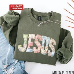 May include: Military green sweatshirt featuring the word "JESUS" in a patchwork design of floral and gingham fabrics. A silver necklace with a pendant is on the sweatshirt. The sweatshirt is paired with blue jeans.