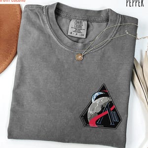 May include: Pepper-colored Comfort Colors t-shirt with a crew neck and a graphic patch featuring a moon and the names Wiseman, Glover, Koch, and Hansen. A silver necklace with a pendant is draped across the shirt.