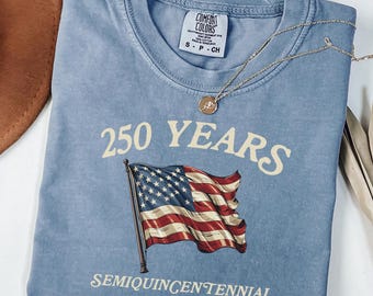 May include: A blue jean colored t-shirt with the text "250 YEARS" and a graphic of the American flag. The shirt also has the text "SEMIQUINCENTENNIAL since 1776". A gold necklace is draped across the shirt.