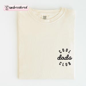 May include: Cream-colored t-shirt with the text "Cool Dads Club" in black script on the lower right. The shirt has a crew neck and is folded neatly. The word "embroidered" is in a pink banner in the upper left.