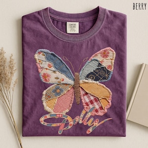 May include: A berry-colored t-shirt featuring a large butterfly graphic made of various patterned fabric patches. The butterfly has a brown stitched body and wings with floral, plaid, and geometric prints. The word Dolly is written below.