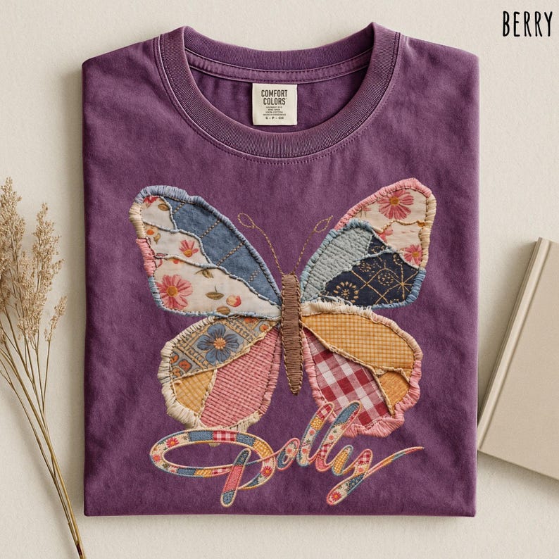 May include: A berry-colored t-shirt featuring a large butterfly graphic made of various patterned fabric patches. The butterfly has a brown stitched body and wings with floral, plaid, and geometric prints. The word Dolly is written below.