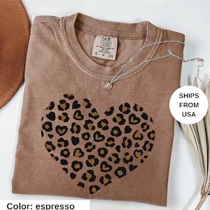 May include: Espresso-colored t-shirt with a leopard print heart design. The shirt has a crew neck and short sleeves. A silver necklace with a small pendant is draped across the shirt. The shirt is labeled "Comfort Colors".