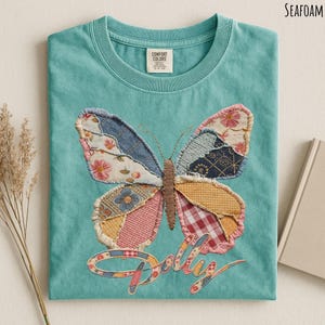 May include: A seafoam green t-shirt featuring a large butterfly graphic made of various fabric patches in floral, plaid, and geometric patterns. The butterfly has a brown stitched body, and the word "Dolly" is written below in a cursive font.