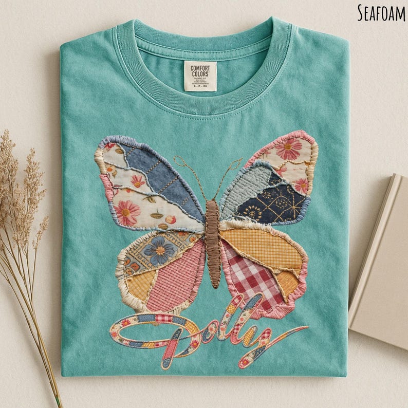May include: A seafoam green t-shirt featuring a large butterfly graphic made of various fabric patches in floral, plaid, and geometric patterns. The butterfly has a brown stitched body, and the word "Dolly" is written below in a cursive font.