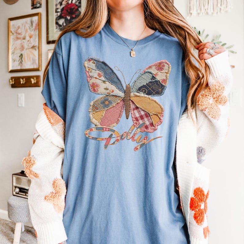 May include: A blue t-shirt featuring a large butterfly graphic composed of various patchwork fabric patterns in shades of pink, yellow, blue, and brown. The word Dolly is printed in a cursive script below the butterfly in a matching patchwork style.