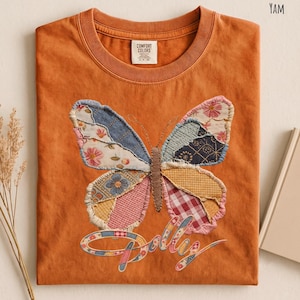 May include: A burnt orange crew neck t-shirt featuring a large butterfly graphic made of various patterned fabric patches. The butterfly has a brown embroidered body and is positioned above the word Dolly in a cursive, multi-colored fabric font.