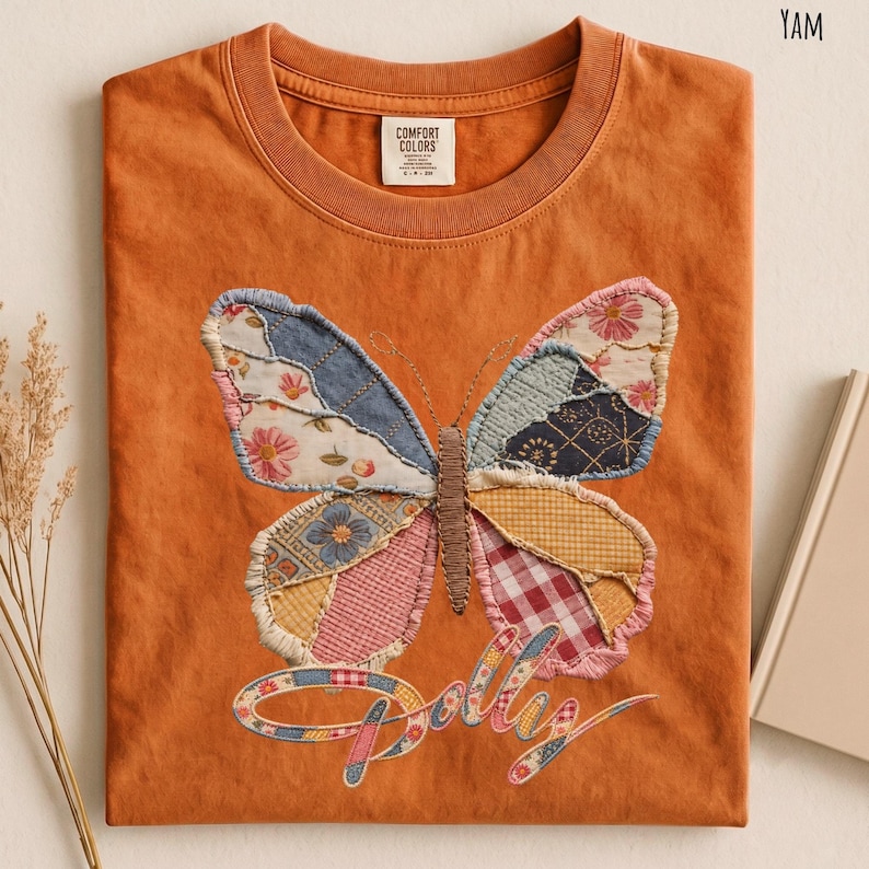 May include: A burnt orange crew neck t-shirt featuring a large butterfly graphic made of various patterned fabric patches. The butterfly has a brown embroidered body and is positioned above the word Dolly in a cursive, multi-colored fabric font.