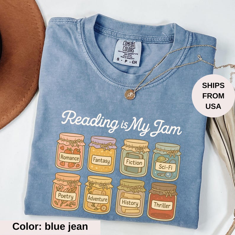 May include: Blue jean Comfort Colors t-shirt with the phrase "Reading is My Jam" and illustrations of jam jars labeled with book genres. A gold necklace is draped across the shirt.