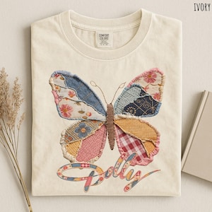 May include: An ivory crew neck t-shirt featuring a large butterfly graphic composed of various fabric patches in blue, pink, yellow, and red floral and plaid patterns. The word "Dolly" is embroidered in a cursive script below the butterfly.
