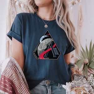 May include: Navy blue t-shirt featuring a black, white, and red graphic design. The design includes a stylized moon, Earth, and the names "WISEMAN", "GLOVER", "KOCH", and "HANSEN". The graphic is within a black triangle.