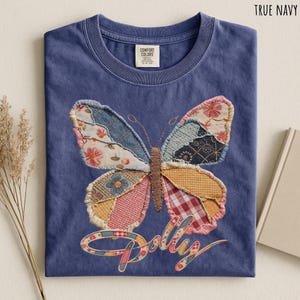 May include: A true navy blue t-shirt featuring a large, central butterfly graphic composed of various patchwork fabric patterns in shades of pink, yellow, blue, and white. The word Dolly is written below the butterfly in a cursive script font.