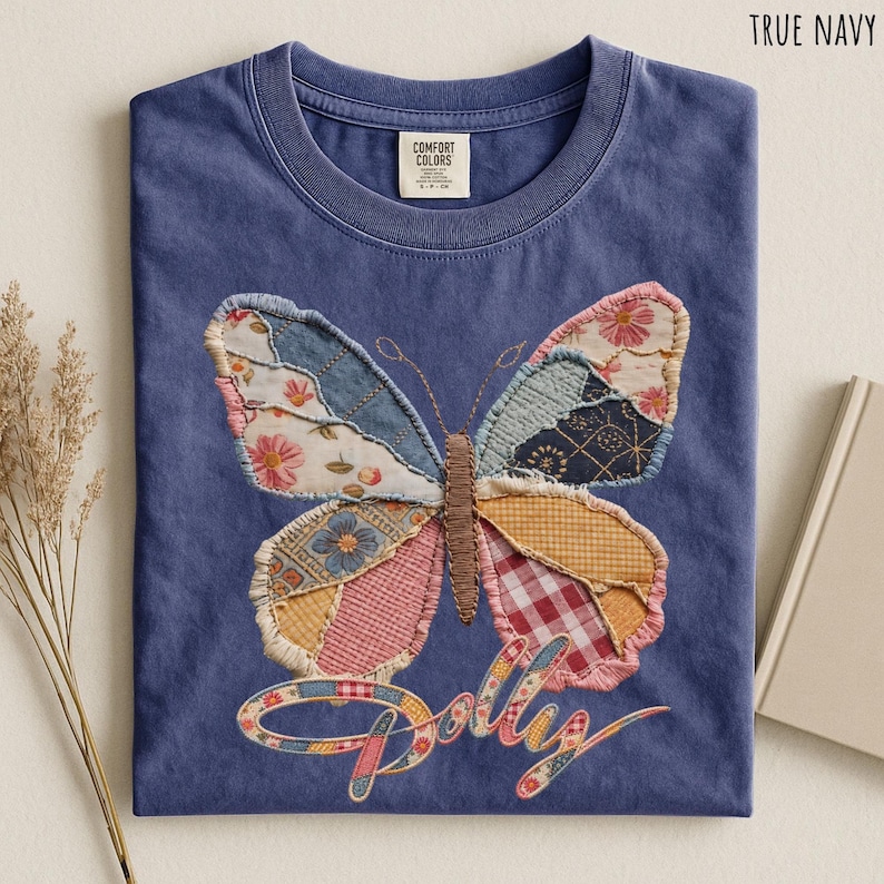 May include: A true navy blue t-shirt featuring a large, central butterfly graphic composed of various patchwork fabric patterns in shades of pink, yellow, blue, and white. The word Dolly is written below the butterfly in a cursive script font.