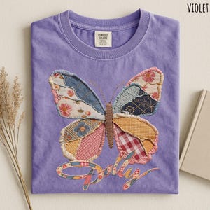 May include: A violet t-shirt featuring a large butterfly graphic composed of various fabric patches in floral, plaid, and geometric patterns. The word Dolly is written in a cursive script below the butterfly, also filled with a patchwork pattern.