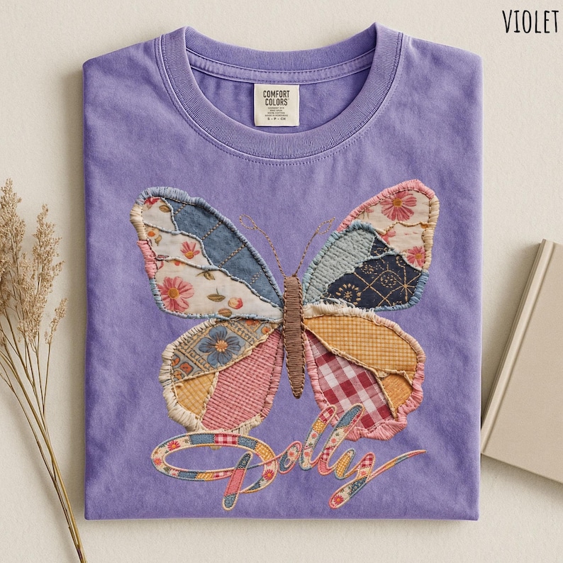 May include: A violet t-shirt featuring a large butterfly graphic composed of various fabric patches in floral, plaid, and geometric patterns. The word Dolly is written in a cursive script below the butterfly, also filled with a patchwork pattern.