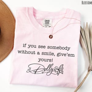 May include: A light pink t-shirt featuring the black printed text, if you see somebody without a smile, give'em yours! and the name Dolly in a cursive script with a butterfly graphic. The shirt has a Comfort Colors brand label at the neck.