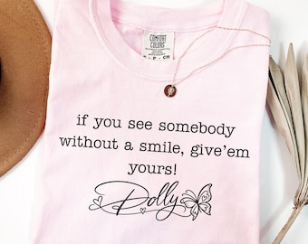 May include: A light pink t-shirt featuring the black printed text, if you see somebody without a smile, give'em yours! and the name Dolly in a cursive script with a butterfly graphic. The shirt has a Comfort Colors brand label at the neck.